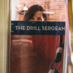 Woman gazes out from behind a poster with the words "The Drill Sergeant".